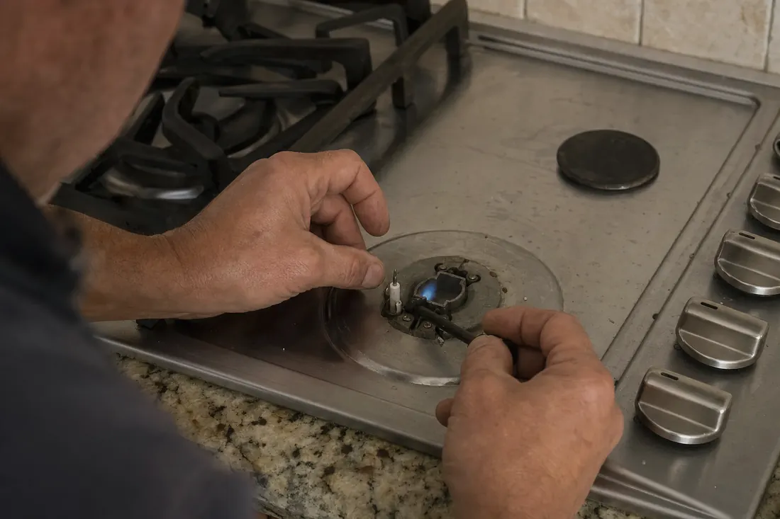 Hands relighting the igniter on a gas cooktop, one burner cap set aside - Range, Cooktop and Wall Oven Repair
