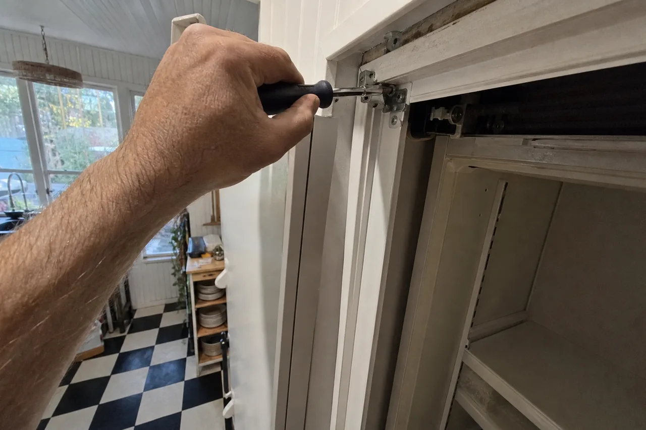 A hand turning a black-handled screwdriver on the top hinge bracket of a tall white built-in column, a checkerboard floor and plate shelves behind - Miele vs Thermador Refrigerator