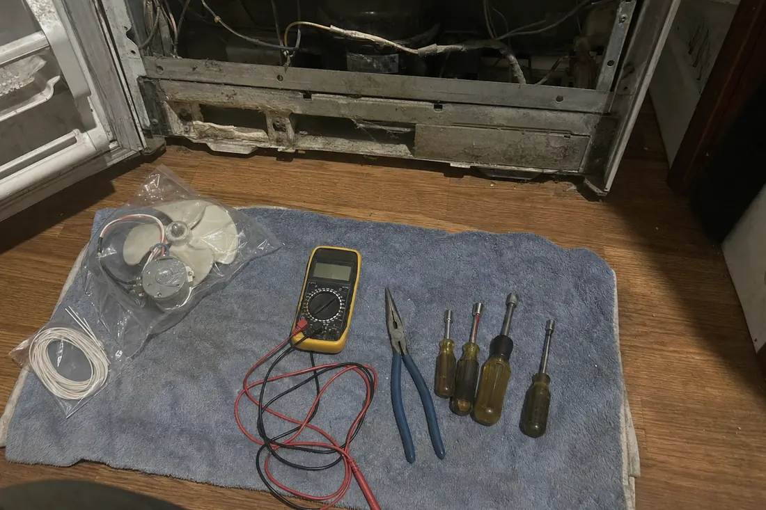 Flat-lay of nut drivers, pliers, a multimeter and a bagged fan motor on a towel, the open refrigerator behind in frame - How a Built-In Ice Maker Fails, and in What Order