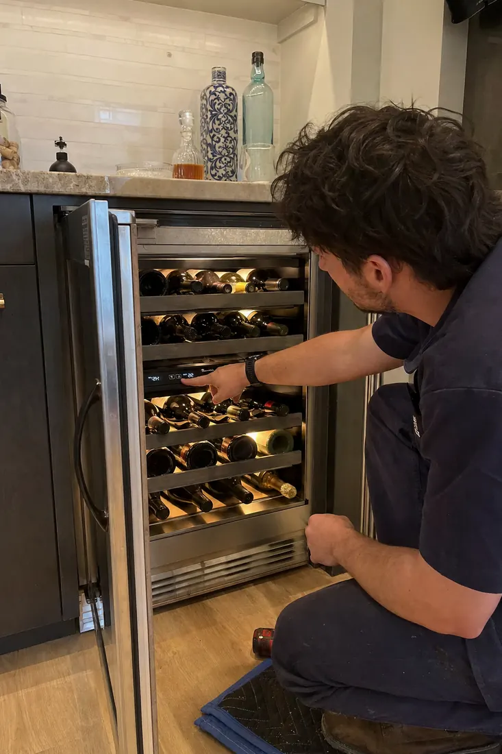 Technician at an open dual-zone wine unit, checking the temperature panel, the open glass door in frame - Guides